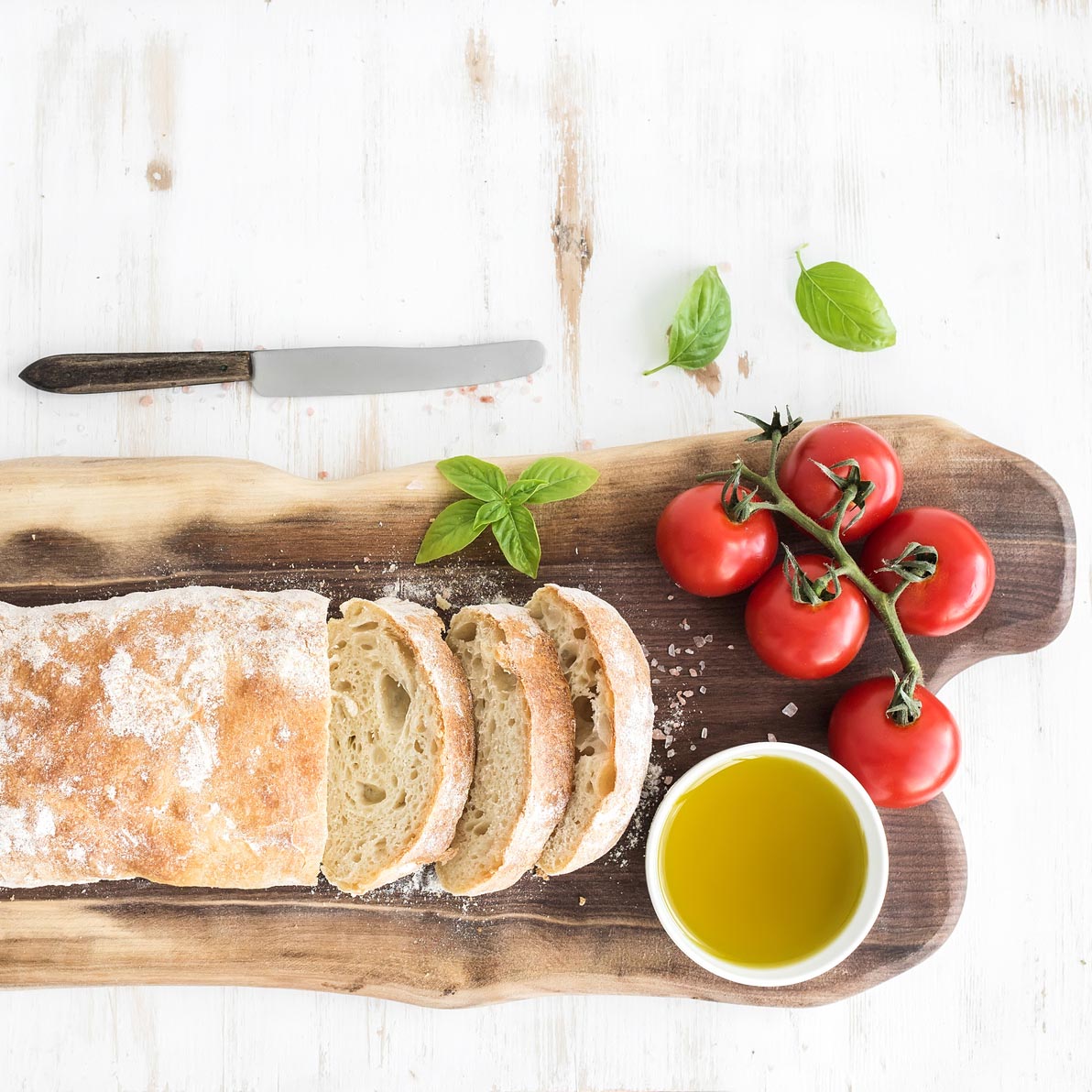 Freshly baked ciabatta bread with cherry-tomatoes, olive oil, basil and salt on walnut wood board over white background, top view, copy space